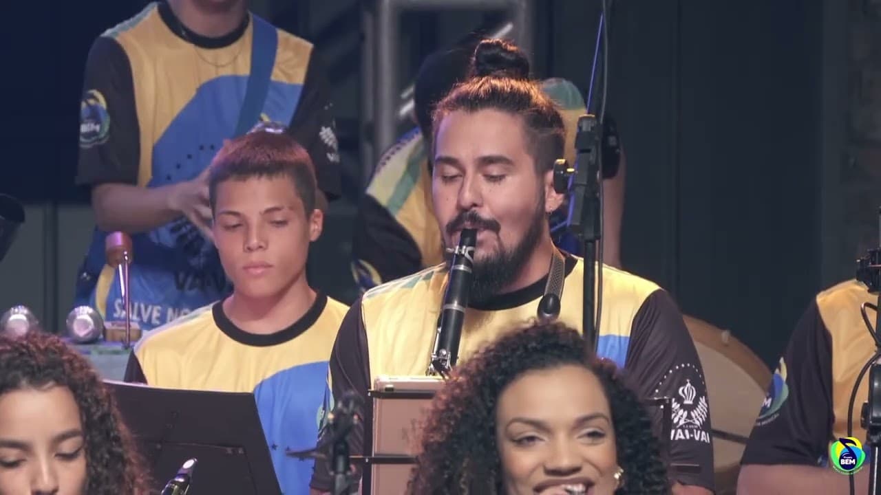Músicos de uniforme amarelo e azul tocando clarinete e percussão, com uma cantora sorrindo em primeiro plano