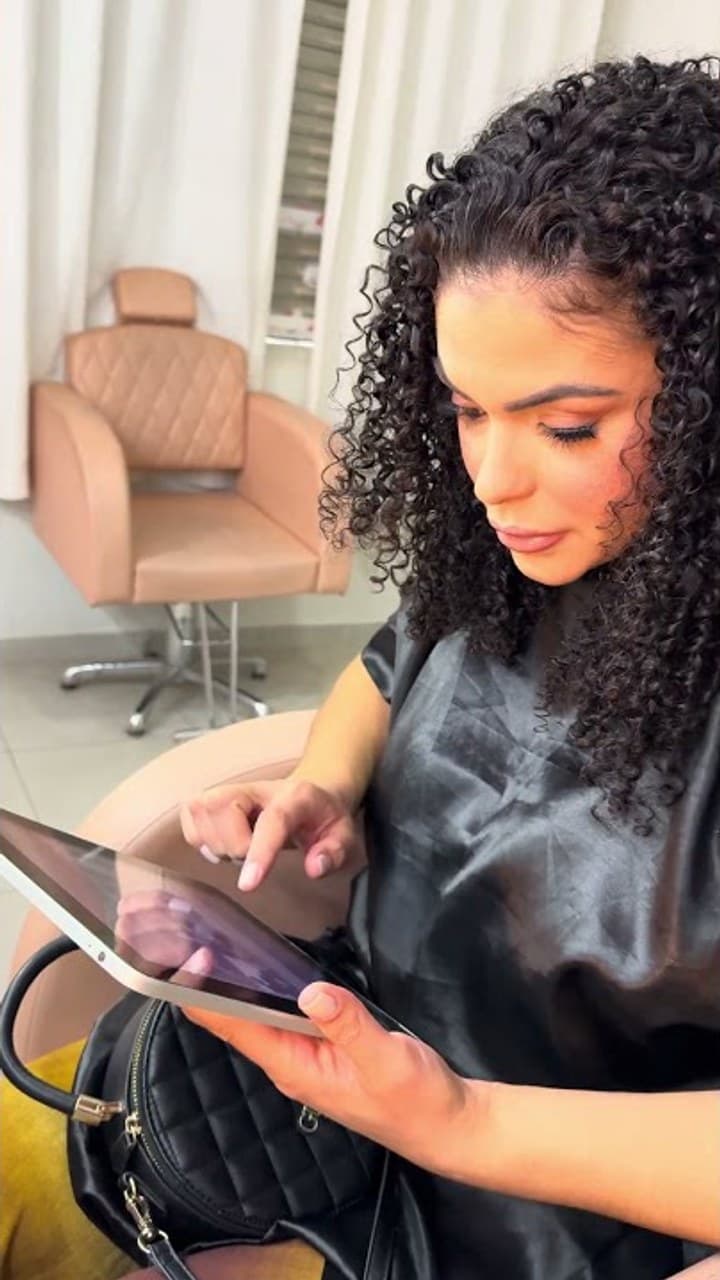 Mulher de cabelo cacheado e capa preta olhando para um tablet, sentada em uma cadeira de salão