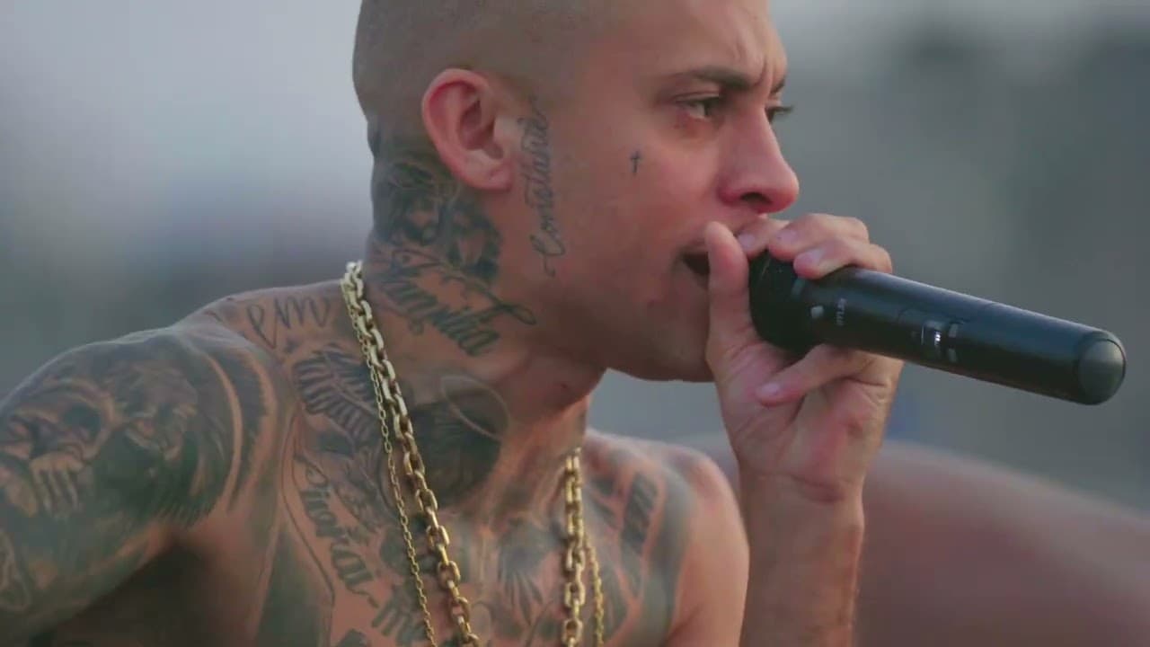 Homem tatuado e sem camisa, com correntes douradas, cantando ao microfone ao ar livre