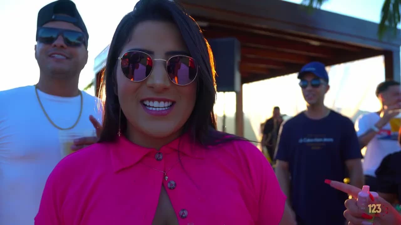 Mulher de óculos escuros redondos e blusa rosa sorrindo em um evento ao ar livre, com pessoas ao fundo