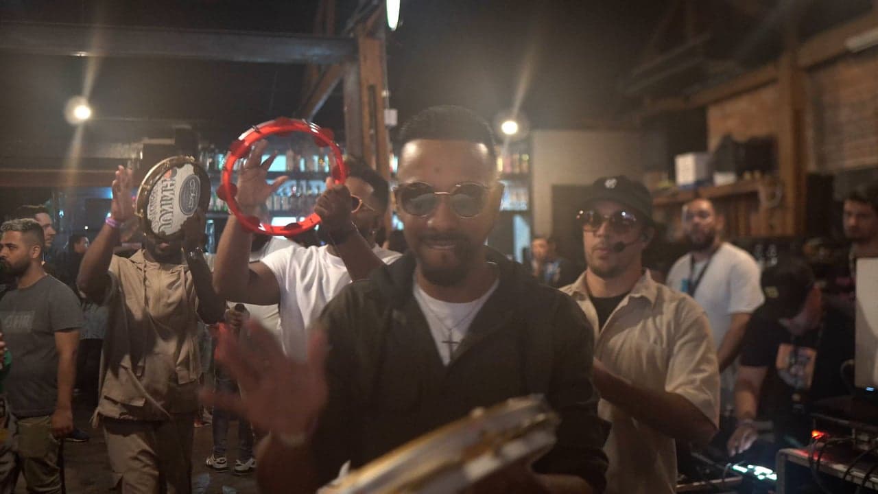 Integrantes do grupo tocando pandeiros em um bar, com um cantor de óculos escuros em primeiro plano