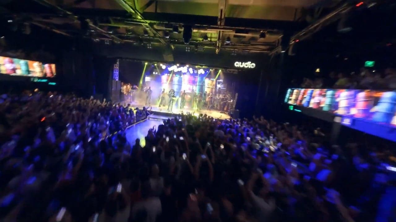 Vista da plateia de braços erguidos diante do palco iluminado, com painéis de LED coloridos dos lados