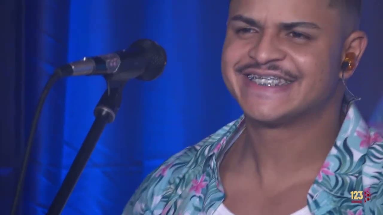Cantor de camisa havaiana sorrindo ao lado do microfone, com cortina azul ao fundo
