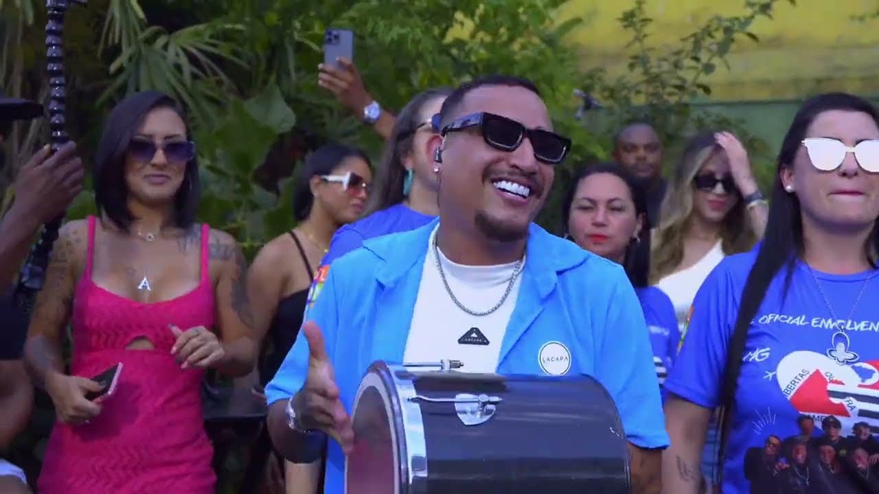 Homem de óculos escuros e camisa azul sorrindo com um instrumento de percussão, cercado por pessoas ao ar livre