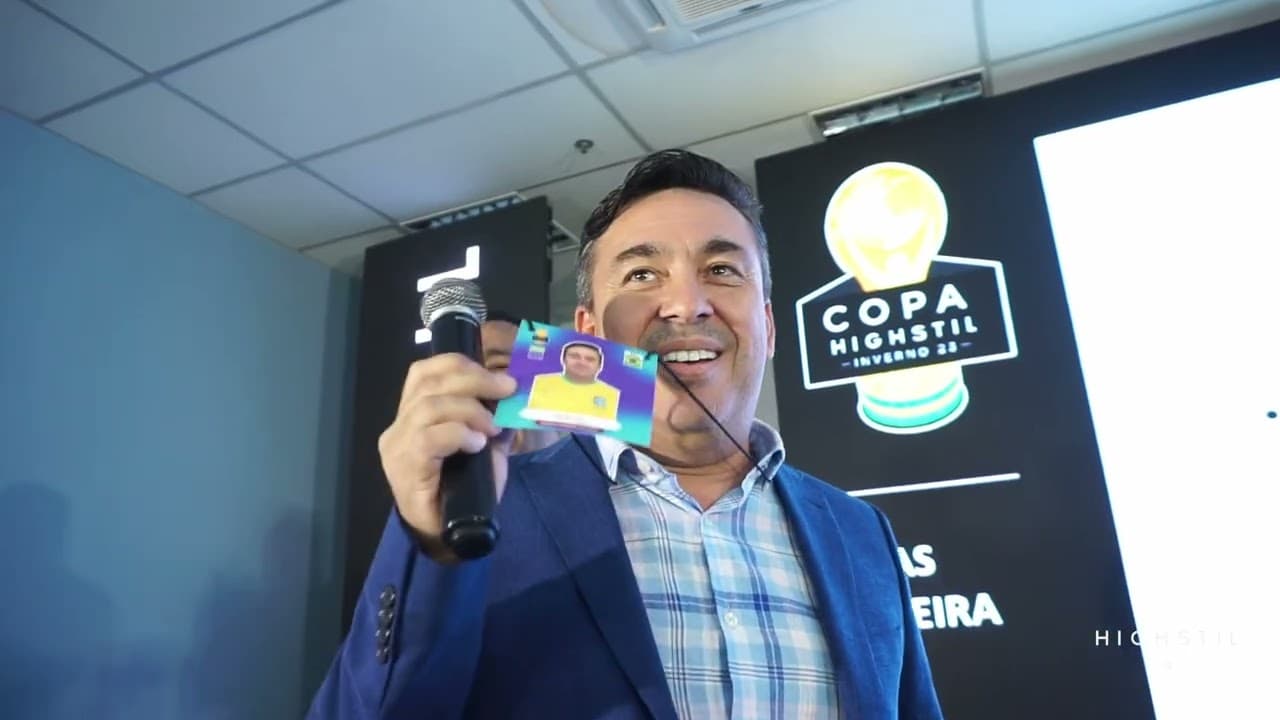 Homem de blazer azul sorrindo com um microfone e uma figurinha na mão, diante de um painel com a logo da Copa Highstil Inverno 23