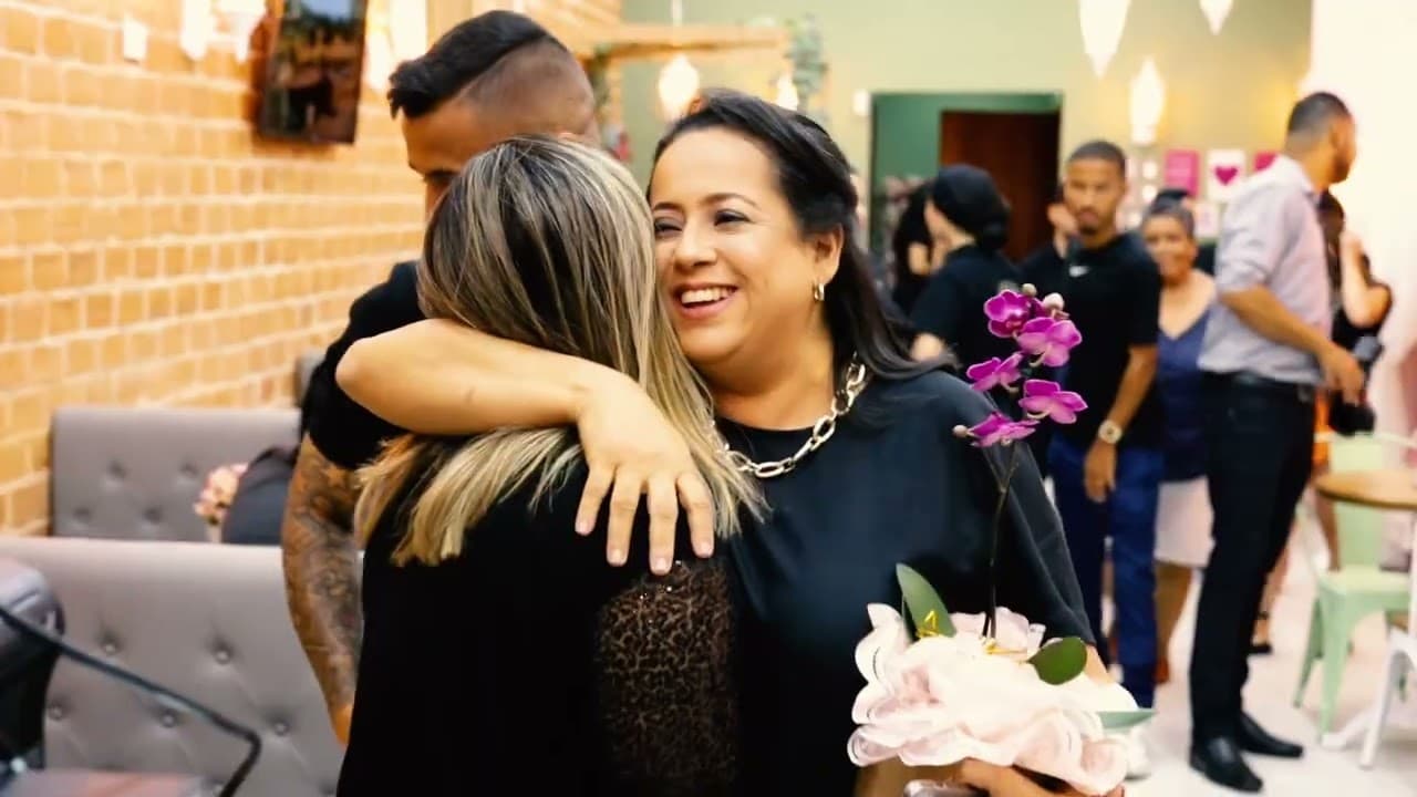 Mulher sorrindo abraçando outra pessoa em uma doceria, com um buquê de flores e pessoas ao fundo