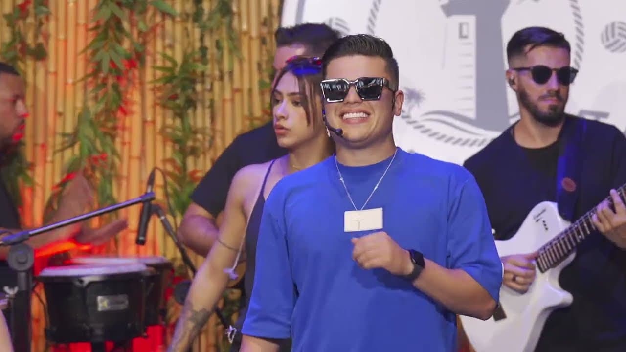 Homem de óculos escuros e camiseta azul sorrindo diante da banda, em um palco com fundo de bambu