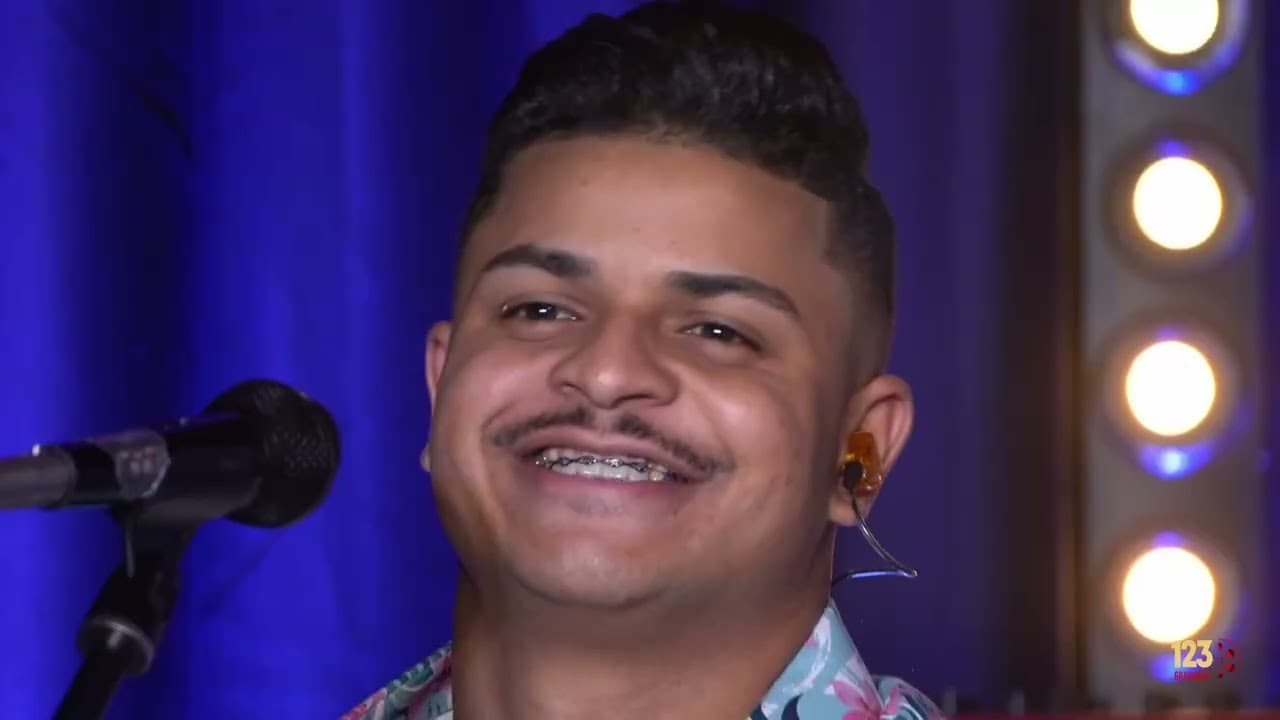 Cantor de camisa estampada sorrindo ao lado do microfone, com cortina azul e refletores ao fundo