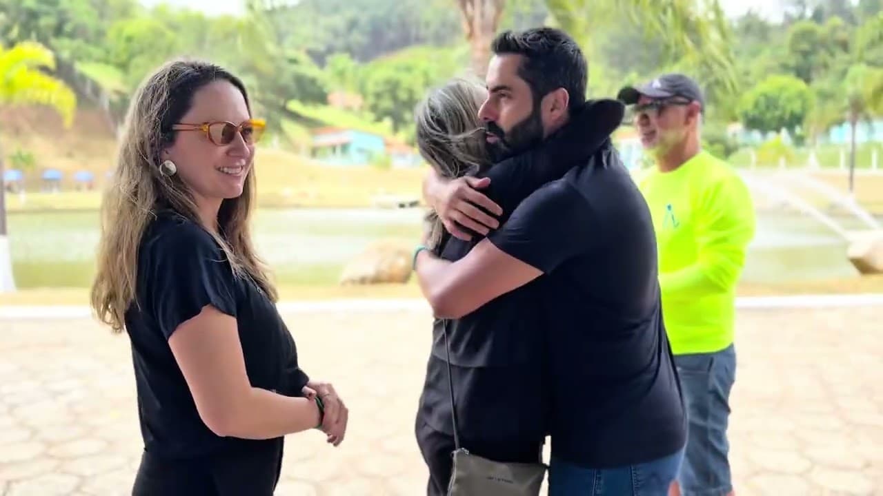 Duas pessoas se abraçando ao ar livre, com uma mulher sorrindo ao lado e um lago ao fundo