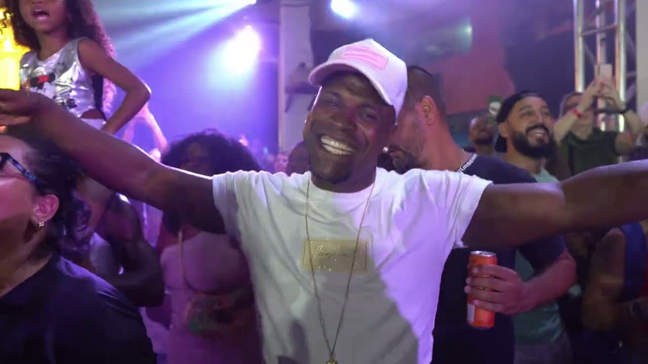 Homem de boné rosa e camiseta branca sorrindo de braços abertos em meio ao público, sob luzes roxas