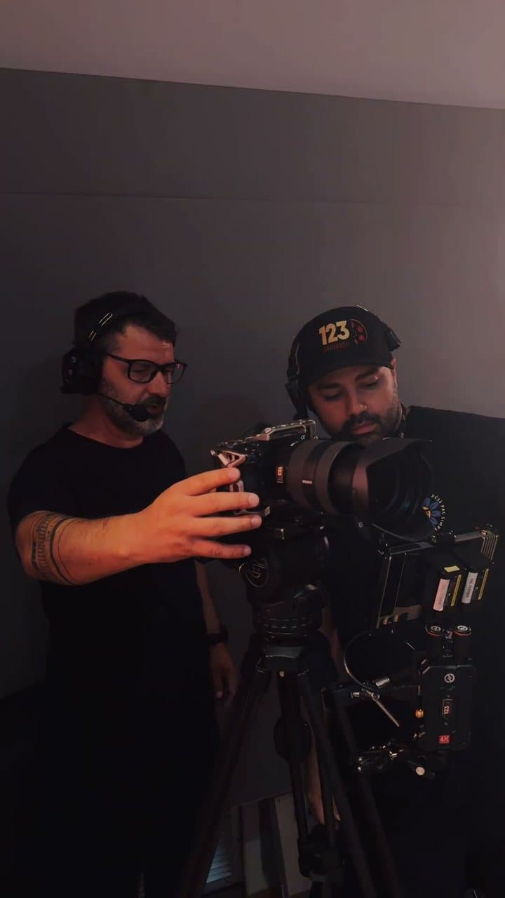 Dois homens de fone com microfone operando uma câmera de cinema sobre tripé, em uma sala escura