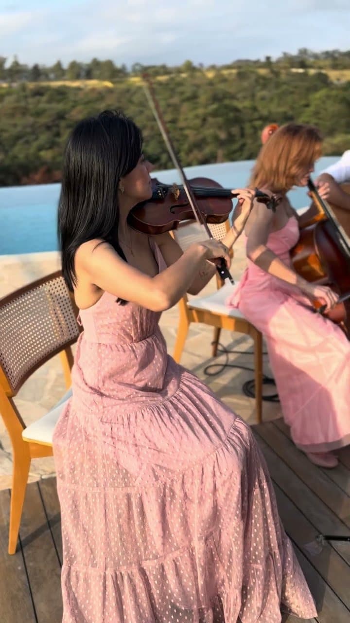 Duas mulheres de vestido rosa tocando violino e violoncelo ao lado de uma piscina, com paisagem de montanhas ao fundo