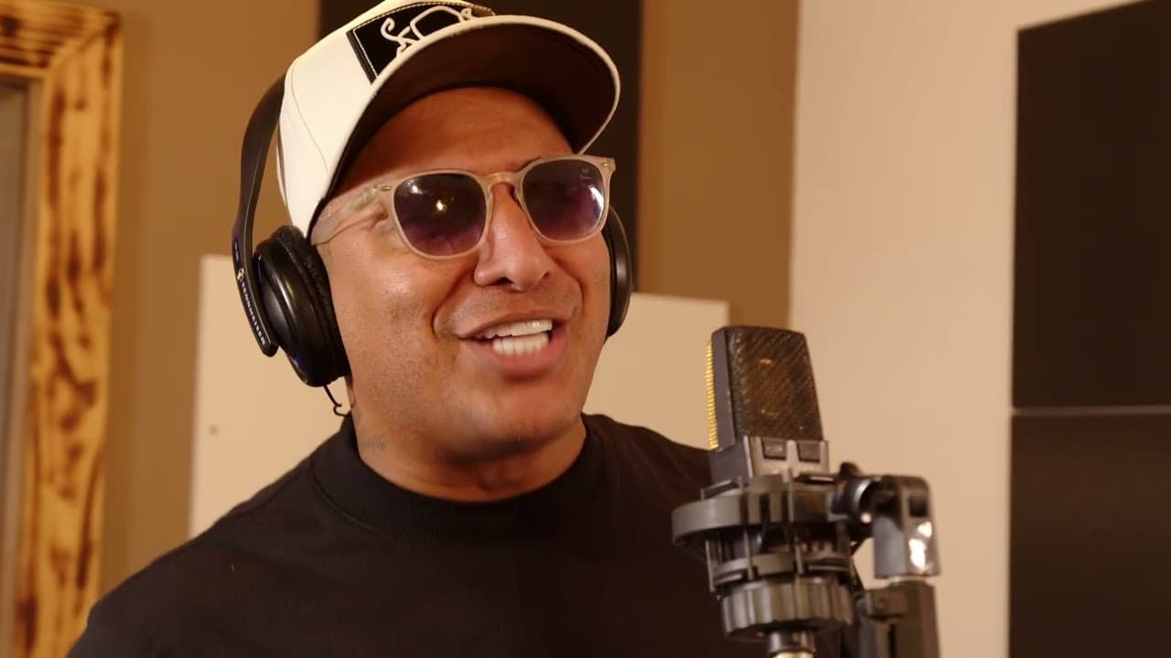 Cantor de boné branco, óculos escuros e fone de ouvido sorrindo diante de um microfone de estúdio