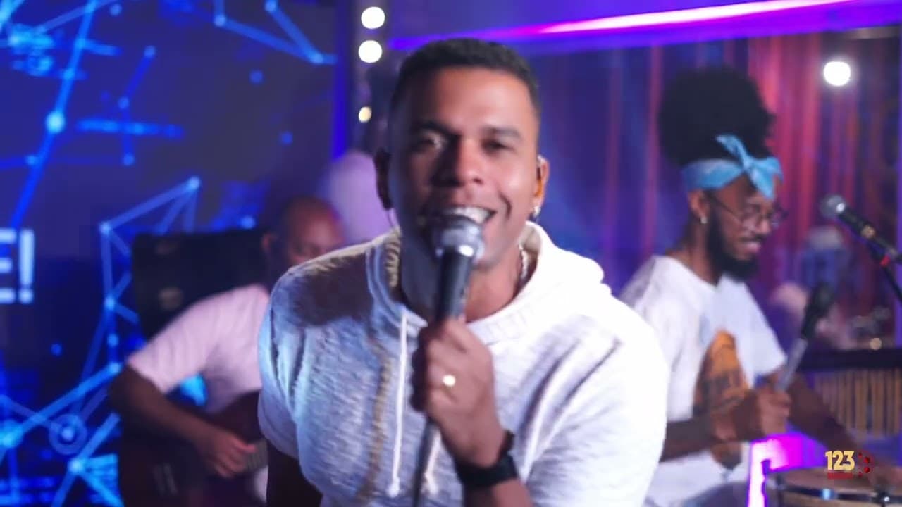 Cantor de moletom branco cantando ao microfone, com músicos tocando violão e percussão e um painel de LED azul ao fundo
