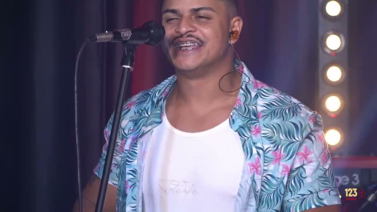 Cantor de camisa havaiana azul e camiseta branca sorrindo ao microfone, com cortina e refletores ao fundo